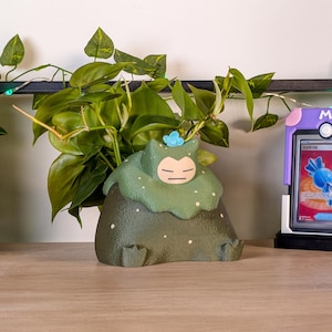 May include: A green and brown ceramic planter shaped like a sleeping cartoon character, with a light blue flower accent. The planter is filled with green leafy plants. A purple card holder with a trading card is visible in the background.
