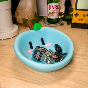 May include: A light blue apple-shaped bowl with a brown stem and green leaf. The bowl features a cartoon face with black eyes and pink cheeks. Inside the bowl sits a silver digital wristwatch with a metal link band, a set of keys, and a black object.