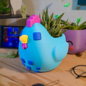 May include: A light blue chicken-shaped planter with a bright pink comb, a yellow beak, and dark blue square accents. The planter holds a green succulent plant. The design features a smooth, matte finish with a rounded body and a wing-like side.