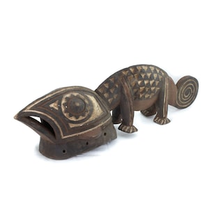 Può includere: Una scultura in legno di un camaleonte con motivi geometrici. Il camaleonte è marrone con accenti dorati e ha una coda arricciata. La scultura fa parte di un pezzo più grande che include una maschera con un grande occhio e motivi geometrici.