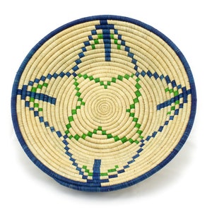 Può includere: Un cesto rotondo intrecciato con un motivo geometrico blu e verde. Il cesto è fatto di materiali naturali e ha un colore paglia naturale.