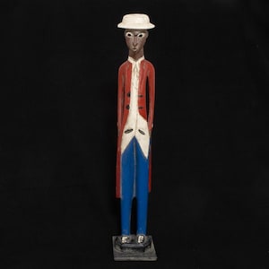 Op de afbeelding: Een houten beeldje van een persoon die een witte hoed, een rood jasje en een blauwe broek draagt. Het beeldje is geschilderd in een volkskunststijl.