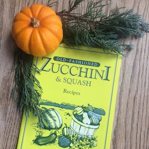 May include: A yellow cookbook with the title "Old-Fashioned Zucchini & Squash Recipes" and the text "Bear Wallow Books" on the cover. A small orange pumpkin sits on top of the book.