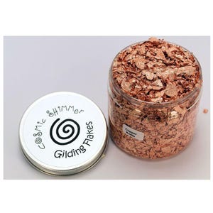 May include: A clear plastic jar filled with copper-colored gilding flakes, labeled "Copper Kettle." Beside it, a small, round tin with a white lid features the text "Cosmic Shimmer Gilding Flakes" and a spiral design.