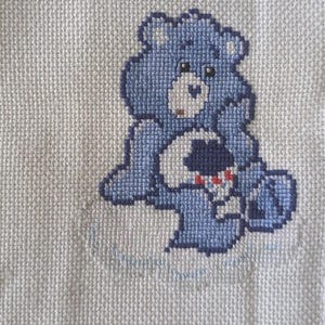 May include: A cross-stitch embroidery on white fabric featuring a light blue bear character with a dark blue nose and a white belly patch. The belly patch displays a dark blue cloud graphic with three small red raindrops falling from it.