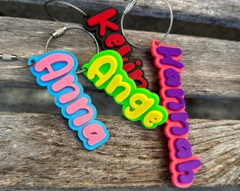May include: Four colorful keychains with names "Anna", "Angie", "Hannah", and "Kevin" written in a bubble font. The keychains are attached to silver metal rings.