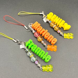 May include: Three colorful phone charms with the names "Genie", "Marlene", and "Frannie" spelled out in 3D letters. Each charm has a gummy bear charm and a string of colorful beads.