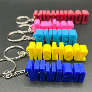 May include: Five colorful keychains with names printed on them. The keychains are in the following order from top to bottom: Hollywood, Ryker, Mstick, Vivian. The keychains are made of a 3D printed plastic material.