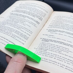 May include: A green page holder with a curved design is placed on a book with French text. The text on the page includes the words "Mettre les verbes entre parenthèses à la forme de participe passé ou d'adjectif verbal selon le sens (observer l'orthographe et la conjugaison)".