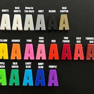 May include: A chart of 18 different colors of plastic letters, each letter is the letter 'A'. The colors are white, silk white, glow in the dark, grey, black, silk copper, yellow, orange, pink, silk pink, red, fire truck, silk red, twinkling red, apple green, silk green, light blue, navy blue, and purple.