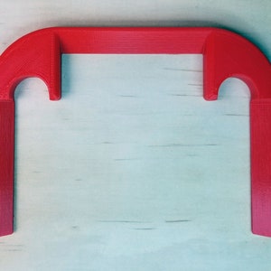 May include: A bright red, 3D-printed object with a rectangular shape and curved ends. The object has a handle-like design with two vertical extensions and a horizontal bar. The item is likely a tool or a decorative piece.