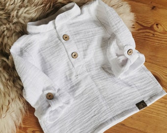 May include: A white long-sleeve shirt with a textured fabric, featuring a mandarin collar, a partial button-down front with two light brown wooden buttons, and roll-up sleeves secured by matching wooden buttons. A small dark tag is on the bottom hem.