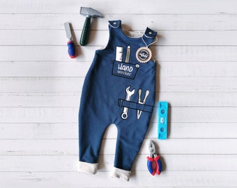 May include: A navy blue sleeveless romper with white cuffs, featuring a graphic print of a pocket containing a ruler and pencil, the text "HAND werker", and a tool belt with a wrench and screwdriver. A round tag is attached to the shoulder strap.