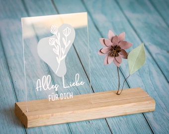May include: A clear rectangular acrylic plaque featuring a white floral line drawing over a light gray organic shape and the white text Alles Liebe FÜR DICH. The plaque is inserted into a rectangular wooden base next to a mauve paper flower.