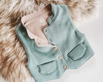 May include: A sage green sleeveless vest with a beige interior lining. The garment features a front button closure with four circular snaps, two decorative front pockets with flaps, and a small circular label printed with the number 62 and EING FADE.