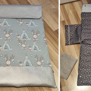 May include: A gray and silver changing mat with a deer and tent pattern. The mat is folded in half and has a silver zipper. The mat is on a wooden floor.