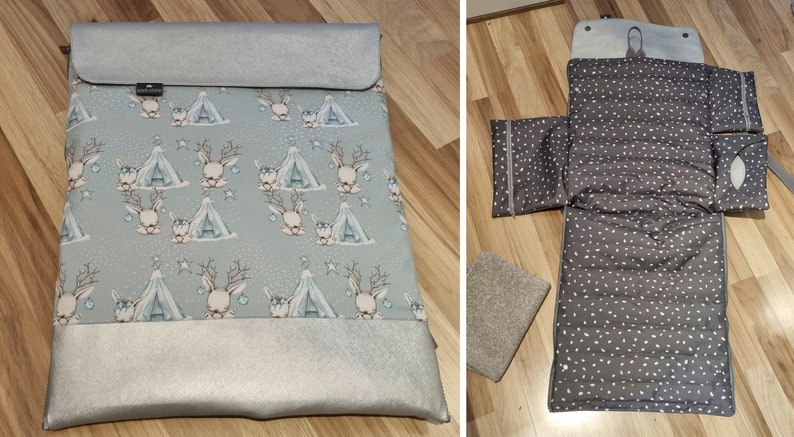 May include: A gray and silver changing mat with a deer and tent pattern. The mat is folded in half and has a silver zipper. The mat is on a wooden floor.