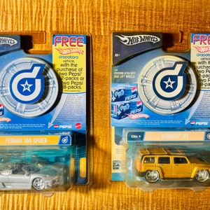 May include: Two Hot Wheels car packaging featuring a red, white, and blue target with a star in the center. The packaging is for a silver Ferrari 360 Spider and a gold Hummer H3. The packaging includes a free car offer with the purchase of Pepsi.