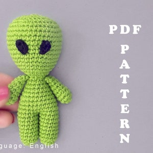 May include: Green crocheted alien amigurumi toy with black eyes. The toy is being held by a hand. The text "PDF PATTERN" is written in white on a gray background.