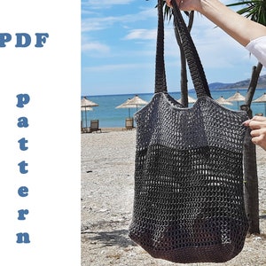 Puede incluir: Una bolsa de mano de crochet gris con una parte superior ancha y abierta y una correa gruesa tejida. La bolsa cuelga de la mano de una persona de pie en una playa.