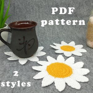 May include: Two crocheted daisy coasters in white and yellow. The coasters are on a gray surface with a mug and a jar of salt. The text "PDF pattern" and "2 styles" is visible in the image.