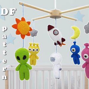 May include: A crocheted baby mobile with a space theme. It features a yellow sun, a white rocket, a blue alien, a green alien, a pink alien, a gray star, and a yellow crescent moon.