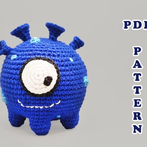 May include: A blue crocheted monster toy with one large white eye, a black mouth, and blue spots. The toy has four legs and three spiky antennas on top of its head. The text "PDF PATTERN" is written in blue letters on the right side of the image.