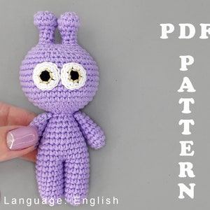 May include: A purple crocheted amigurumi alien toy with white eyes and black pupils. The toy is being held by a hand. The text "PDF PATTERN" is written in white letters on a gray background.