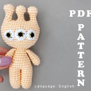May include: Crochet pattern PDF for a three-eyed amigurumi creature with a peach-colored body and white eyes. The pattern is written in English.