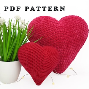 May include: Two red heart-shaped crochet pillows of different sizes. The larger pillow has a plush texture. A small white potted plant with green leaves and white flowers is to the left. The text "PDF PATTERN" is at the top.