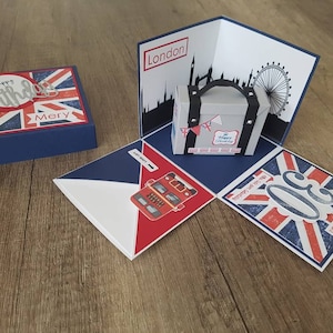 Könnte beinhalten: Eine blaue und rote Box mit einem Union-Jack-Design. Die Box ist geöffnet und zeigt eine Pop-up-Karte mit einem Koffer, einem roten Doppeldeckerbus und einer Londoner Skyline. Die Karte enthält auch den Text "Happy Birthday" und "London".