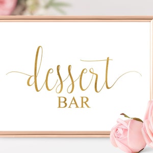 May include: A gold sign with the text "dessert bar" in a cursive font, framed in a rose gold frame. The sign is sitting on a white surface with pink roses in the background.
