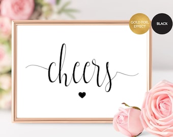 May include: A printable sign with the word "cheers" in a cursive font with a small heart underneath. The sign is in a gold frame and is sitting on a table with pink flowers.