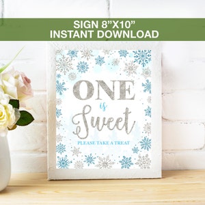 Può includere: Un cartello bianco con la scritta "ONE is Sweet" in lettere glitterate argentate, circondato da fiocchi di neve argentati e blu. Il cartello misura 20,3 cm x 25,4 cm e include la scritta "PLEASE TAKE A TREAT".