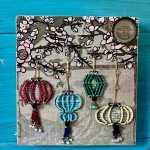May include: A square decorative wall art piece featuring a printed background of cherry blossom branches and four hanging lantern ornaments. The lanterns are crafted from beads in red, light blue, green, and cream, each with a dangling tassel.