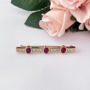 May include: A gold-tone bar brooch featuring three oval-shaped deep red stones set in the centre, alternating with rows of small clear white crystals embedded along the horizontal metal frame, creating a linear and symmetrical decorative accessory.
