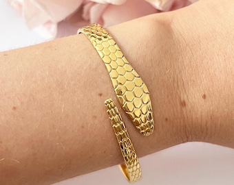 Puede incluir: Un brazalete de serpiente dorado con un diseño texturizado en forma de panal. El brazalete está abierto y se envuelve alrededor de la muñeca.