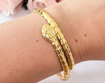 Puede incluir: Una pulsera de serpiente dorada con un diseño detallado. La cabeza de la serpiente presenta dos piedras claras y brillantes como ojos. La pulsera se envuelve alrededor de la muñeca, mostrando un patrón texturizado en forma de escamas.