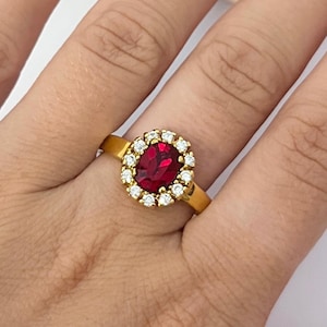 May include: A gold ring featuring a central oval-cut deep red gemstone surrounded by a halo of small, round, clear white stones. The band is a solid, polished gold metal, providing a bright contrast to the vibrant red center stone and the surrounding clear stones.