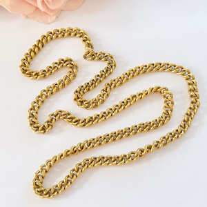 May include: A gold chain necklace with a curb link style. The chain is lying on a white surface.