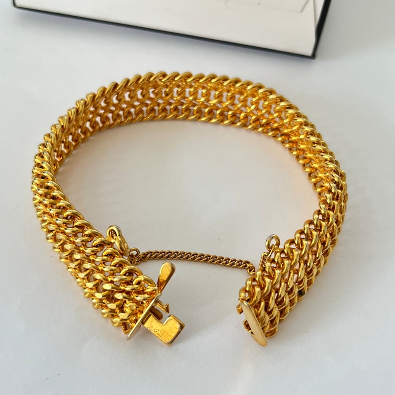 Puede incluir: Un brazalete de cadena de oro con una doble fila de eslabones entrelazados. El brazalete tiene un cierre de broche.