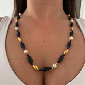 Può includere: Una collana di perline nera con accenti dorati e color crema. La collana presenta perline nere allungate, intervallate da perline ovali color oro e perline di perle color crema. La collana è esposta su uno sfondo bianco.