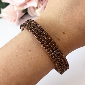 May include: A bracelet featuring four rows of small, round, brown rhinestones set into a light tan band. The accessory wraps around the wrist, displaying a uniform grid of sparkling, dark amber-coloured stones that cover the entire outer surface.