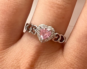 May include: A silver ring with a pink heart-shaped gemstone surrounded by smaller clear gemstones. The ring features a delicate design of interlocking hearts.