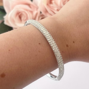 Könnte beinhalten: Ein silberfarbenes Armband mit mehreren Reihen klarer, funkelnder Kristalle. Das gebogene Armband umschließt das Handgelenk und fängt das Licht ein. Im Hintergrund sind zarte rosa Blumen zu sehen.