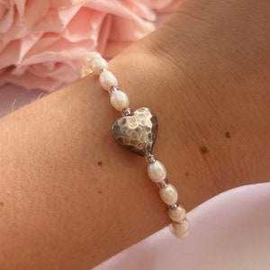 Könnte beinhalten: Ein Armband aus weißen Perlen mit einem gehämmerten silbernen Herz-Anhänger. Das Armband befindet sich an einem Handgelenk.