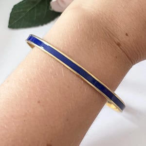 Puede incluir: Un brazalete dorado con una franja central de esmalte azul brillante. El brazalete tiene un diseño sencillo y elegante, perfecto para el uso diario. El brazalete se muestra en el brazo de una persona.