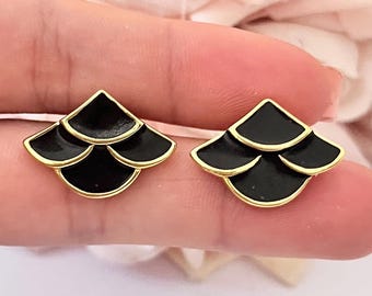 May include: A pair of black and gold stud earrings. Each earring features a fan-shaped design with three overlapping black enamel sections, outlined in gold. The earrings are held up against a soft pink background.