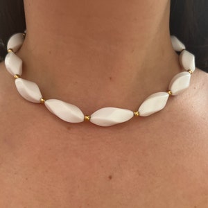 Könnte beinhalten: Eine weiße Keramik-Halskette mit goldenen Akzenten. Die Halskette besteht aus ovalen Perlen mit einem facettierten Design.