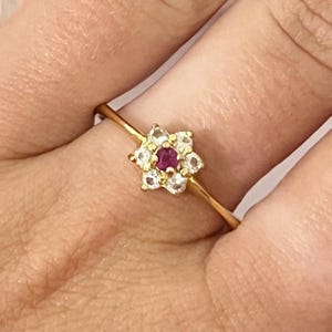 May include: A gold ring featuring a red gemstone surrounded by smaller white gemstones, creating a flower design.